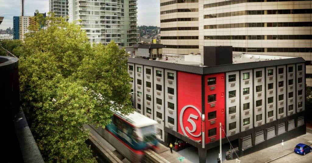 Urban scene with a red building marked with the number 5, surrounded by tall modern buildings. A blurred motion of a tram is seen in front.