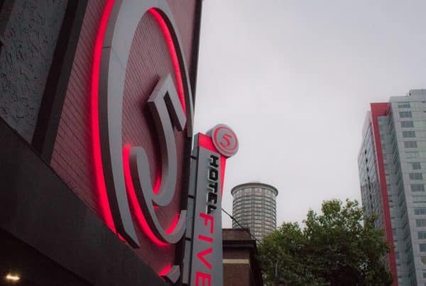 Close-up of a building featuring a red neon "5" logo and "Hotel Five" sign, against a backdrop of overcast sky and surrounding urban structures.