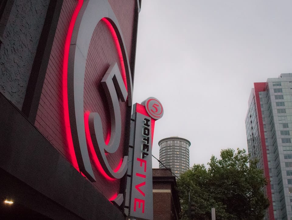 Close-up of a building featuring a red neon "5" logo and "Hotel Five" sign, against a backdrop of overcast sky and surrounding urban structures.