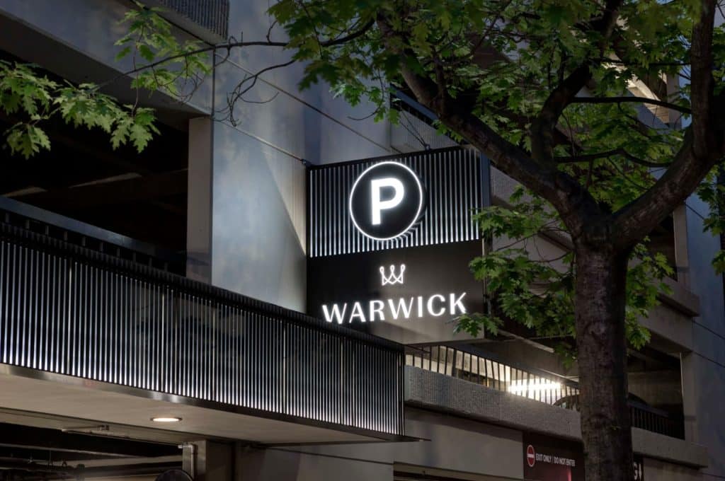 Sign reading "Warwick" with a parking symbol on a building facade, next to a tree.