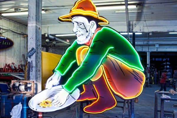 Neon sign of a man in a hat crouching and panning for gold, displayed in an industrial space.