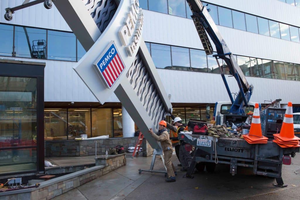 Workers install a large sign on a building. A crane lifts the sign, while two workers guide it. Traffic cones and construction materials are visible nearby.