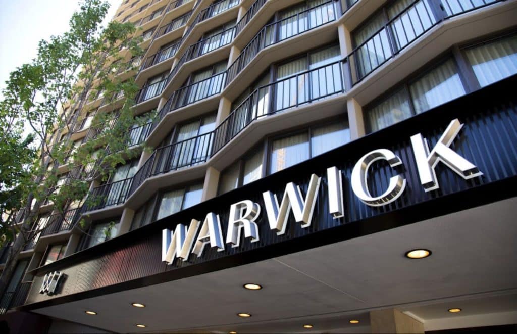 Exterior of the Warwick Hotel with large signage under a high-rise building and adjacent trees.
