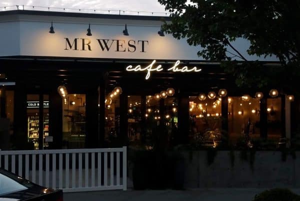 Exterior view of Mr. West cafe bar with illuminated string lights and a dark ambiance. A tree partially frames the right side.