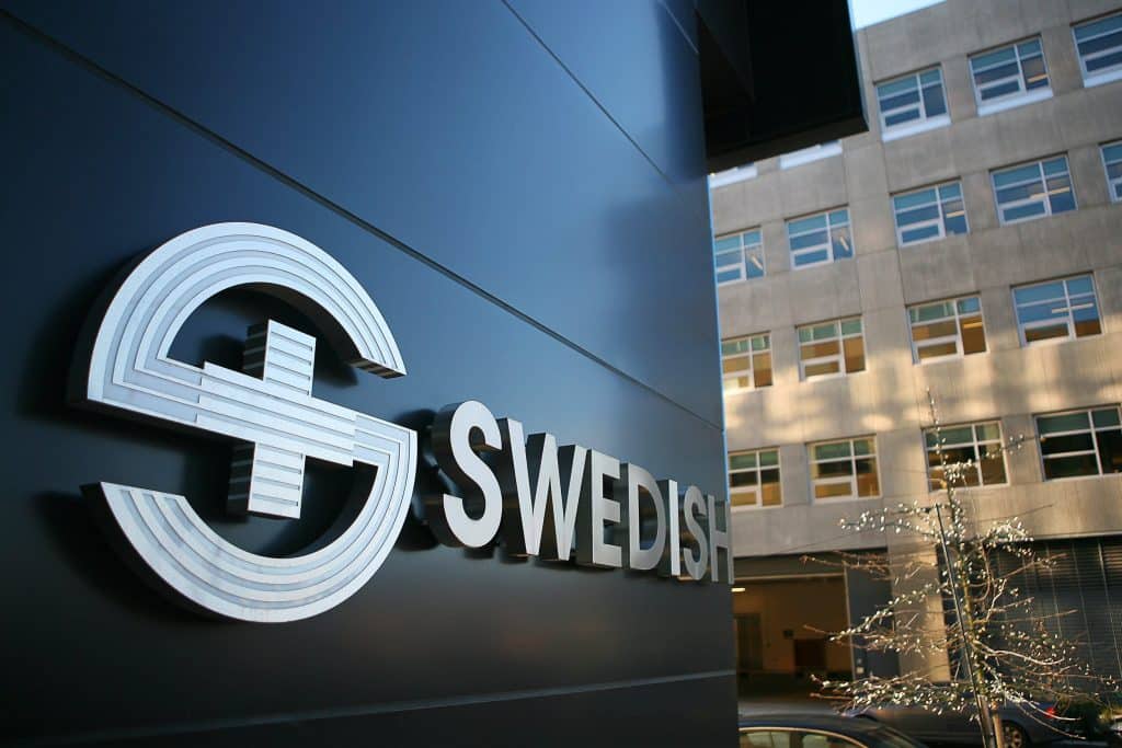 Sign with a stylized "S" symbol and the word "SWEDISH" on a building exterior, with another building and a lit tree in the background. Sign with a stylized "S" symbol and the word "SWEDISH" on a building exterior, with another building and a lit tree in the background.