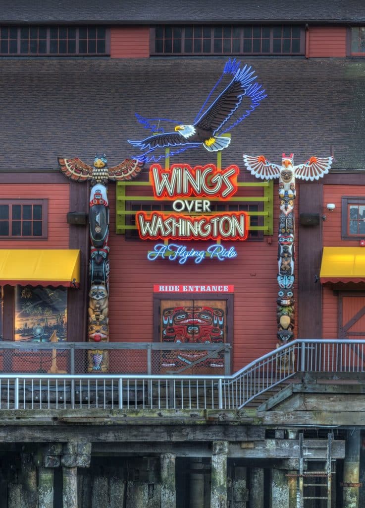 Entrance to "Wings Over Washington" ride featuring neon eagle sign and totem poles on a wooden building with yellow awnings. Entrance to "Wings Over Washington" ride featuring neon eagle sign and totem poles on a wooden building with yellow awnings.