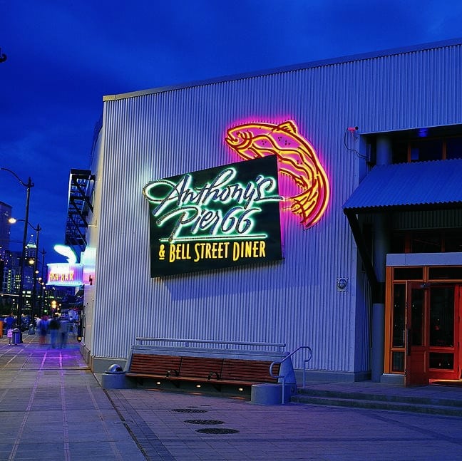 Neon sign on a corrugated metal building reads "Anthony's Pier 66 & Bell Street Diner" with a stylized fish design, shot during dusk, with a cityscape in the background. Neon sign on a corrugated metal building reads "Anthony's Pier 66 & Bell Street Diner" with a stylized fish design, shot during dusk, with a cityscape in the background.