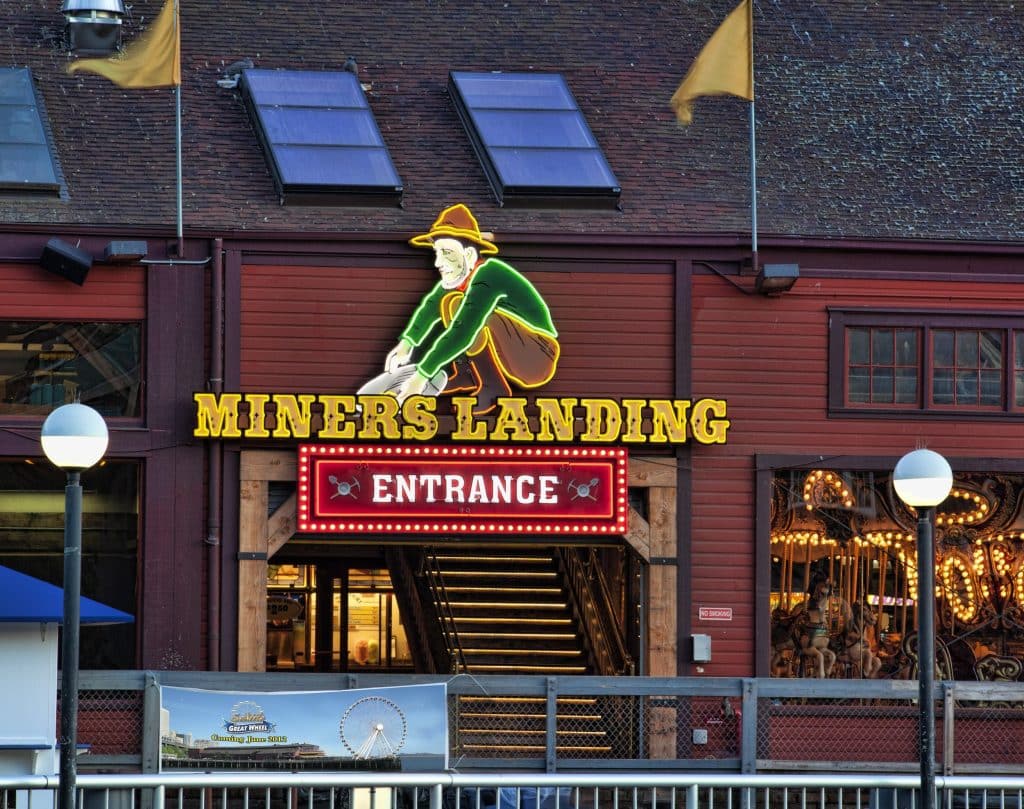 The entrance to Miners Landing, featuring a neon sign with a miner figure above a red "Entrance" sign. The building has wooden siding and two flags on the roof. The entrance to Miners Landing, featuring a neon sign with a miner figure above a red "Entrance" sign. The building has wooden siding and two flags on the roof.