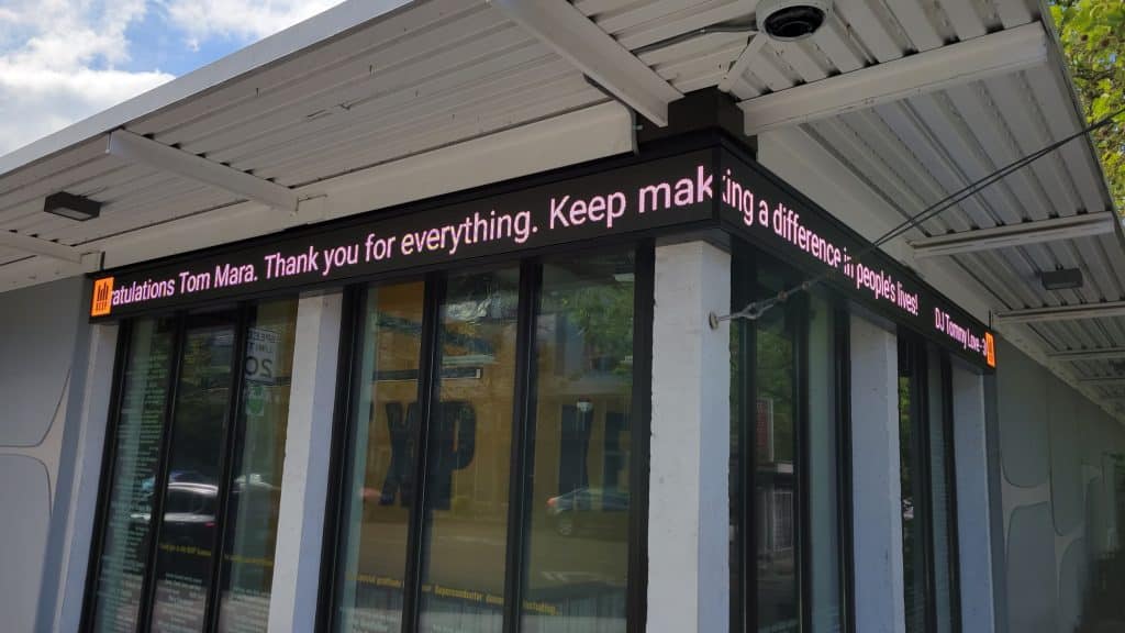 A building corner with a digital marquee reads: "Congratulations Tom Mara. Thank you for everything. Keep making a difference.