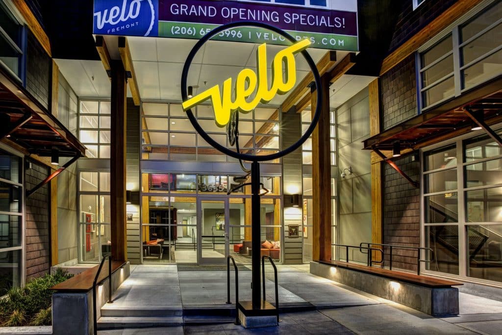 Front entrance of a building with a large circular sign displaying the word "velo." A banner above reads "Grand Opening Specials." The entrance is glass with visible interior seating.
