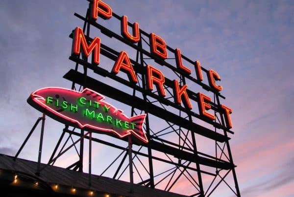 Seattle's Neon illuminates the twilight sky with glowing signs reading "Public Market" and "City Fish Market" atop a bustling building, capturing the city's vibrant charm.