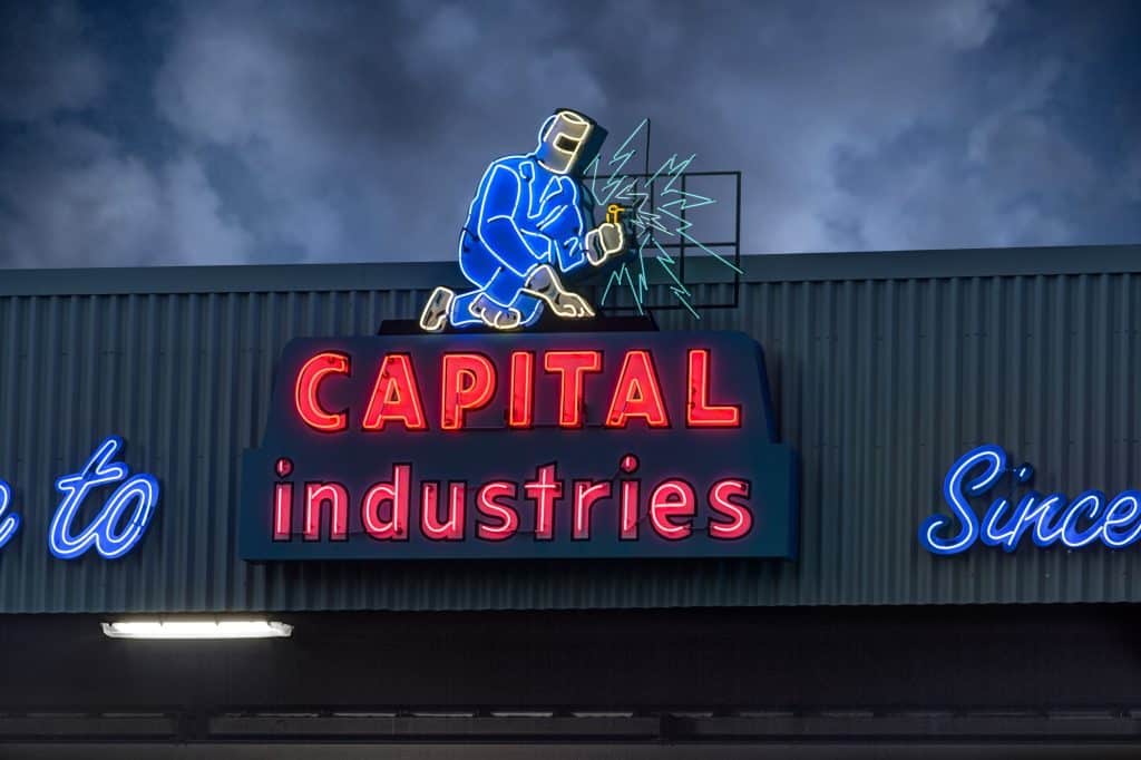 Neon sign depicting a welder above the words "CAPITAL industries" in red and blue on an industrial building.