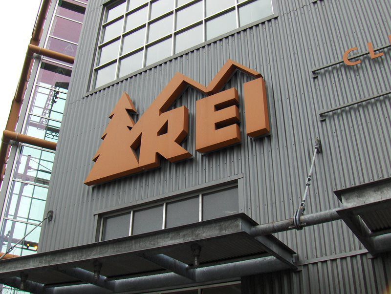 Exterior view of a building with a large REI sign, featuring a logo with a tree and mountain silhouette. Exterior view of a building with a large REI sign, featuring a logo with a tree and mountain silhouette.