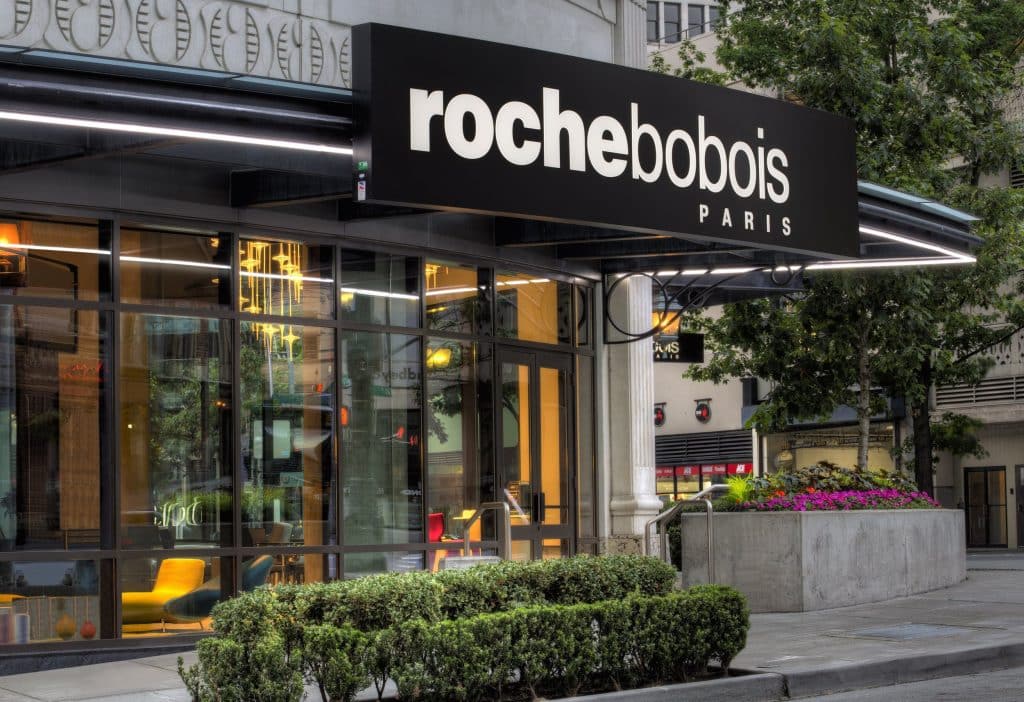 Storefront of Roche Bobois Paris with large glass windows and a black sign. Green shrubs are in front, and colorful furniture is visible inside. Storefront of Roche Bobois Paris with large glass windows and a black sign. Green shrubs are in front, and colorful furniture is visible inside.