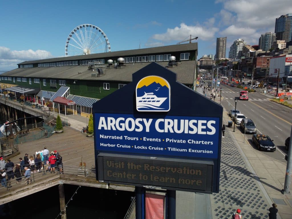 Sign for Argosy Cruises in a waterfront area, with a Ferris wheel, building, and busy street in the background.