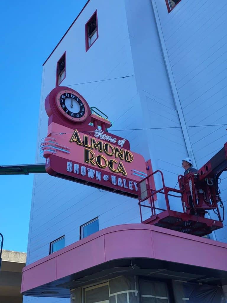A worker in a lift installs a large pink sign saying "Home of Almond Roca, Brown & Haley" on a building, showcasing the sweet legacy celebrated by Almond Roca King across Seattle.