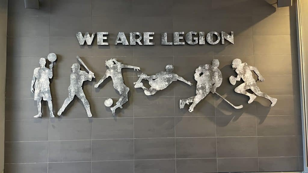 Metallic figures of athletes in various sports poses are displayed on a wall under the text "WE ARE LEGION.