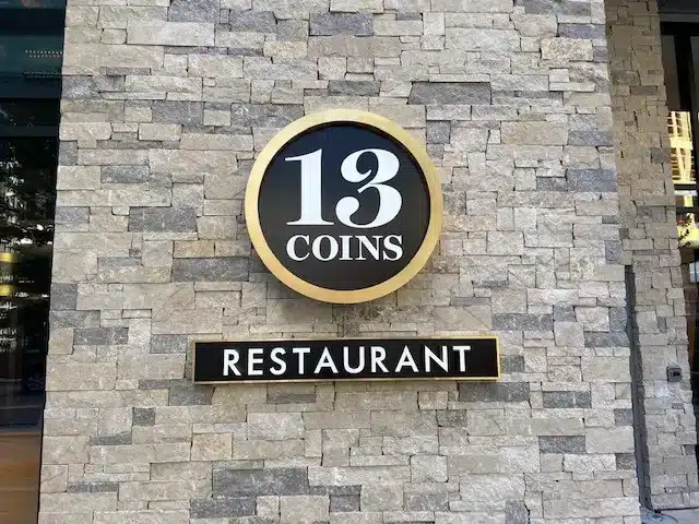 Sign on a brick wall for "13 Coins Restaurant" with a circular logo above the text.