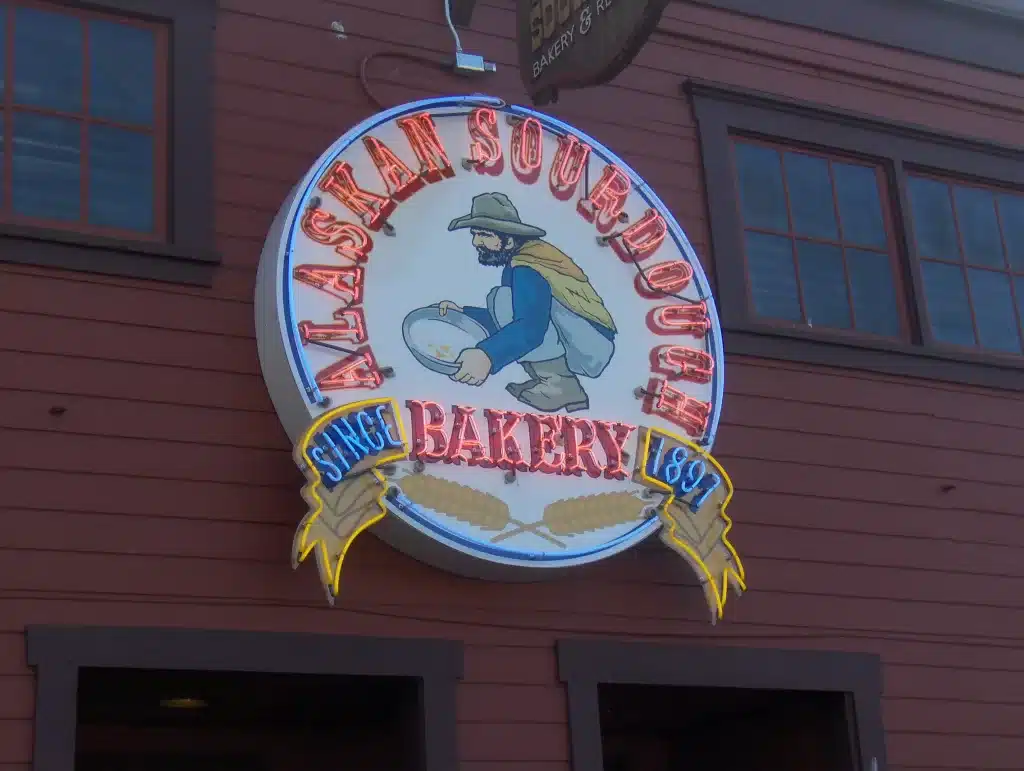 Round sign for "Alaskan Sourdough Bakery" featuring an image of a prospector with a pan, mounted on a red wooden building.