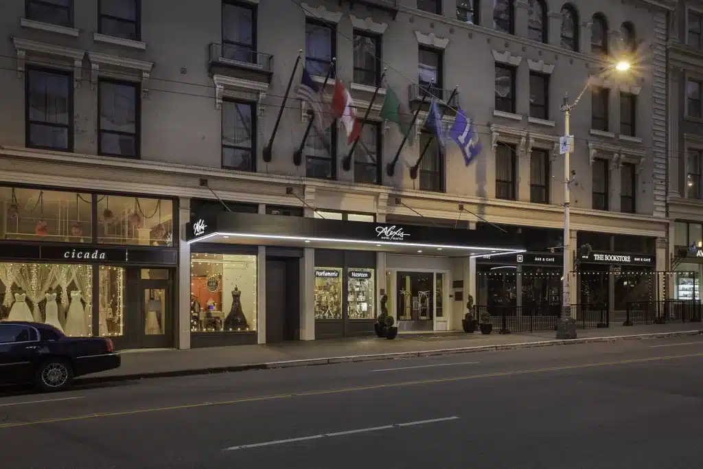 Street view of a large building with multiple storefronts, including Cicada and Teddy's on the Line. Six flags are displayed above the awnings. A car is parked on the street at dusk.
