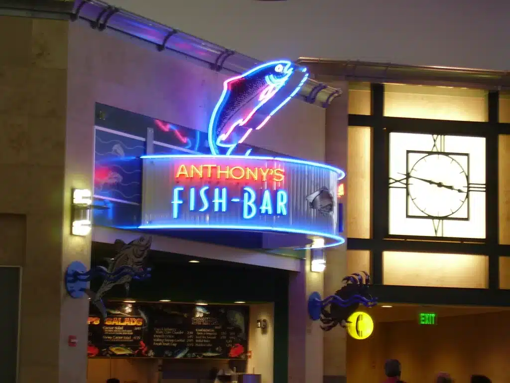 Neon sign reading "Anthony's Fish Bar" with a fish design, located indoors near a clock and illuminated exit sign.