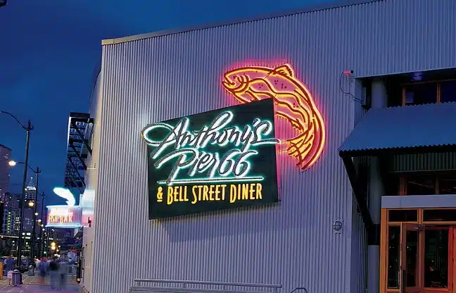 Neon sign for Anthony's Pier 66 & Bell Street Diner on a corrugated metal wall, featuring a fish outline, against a cityscape background at dusk.