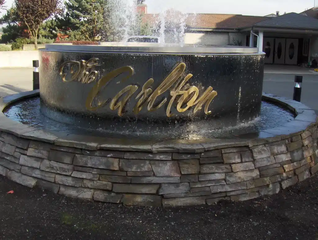 Stone fountain with water flowing over the edge, displaying metallic letters spelling "The Carlton." Surrounded by a stone wall, located outdoors under a clear sky.