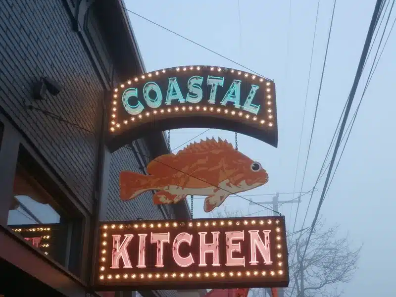 A lit sign displays a fish with "Coastal Kitchen" text below, attached to a building.