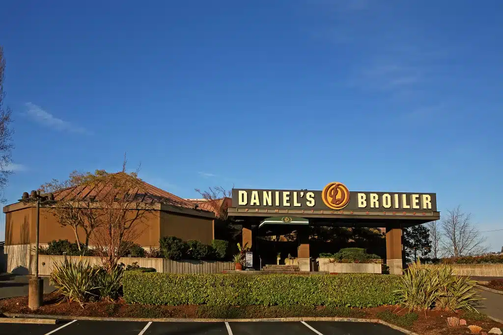 Front exterior of Daniel's Broiler, a restaurant with a large sign and surrounding greenery, under a clear blue sky.