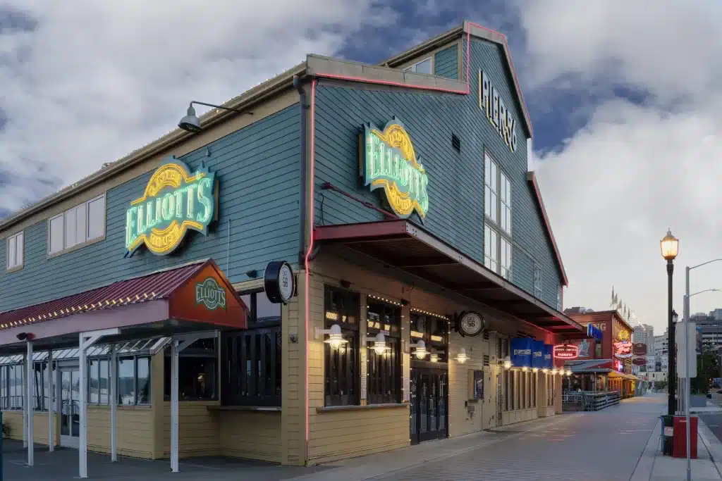 A waterfront restaurant with a sign reading "Elliott's" is located along a paved walkway, under a cloudy sky.