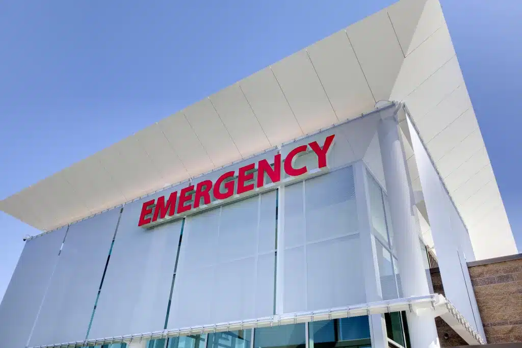 Modern hospital building with a prominent red "Emergency" sign on the facade, under a clear blue sky.