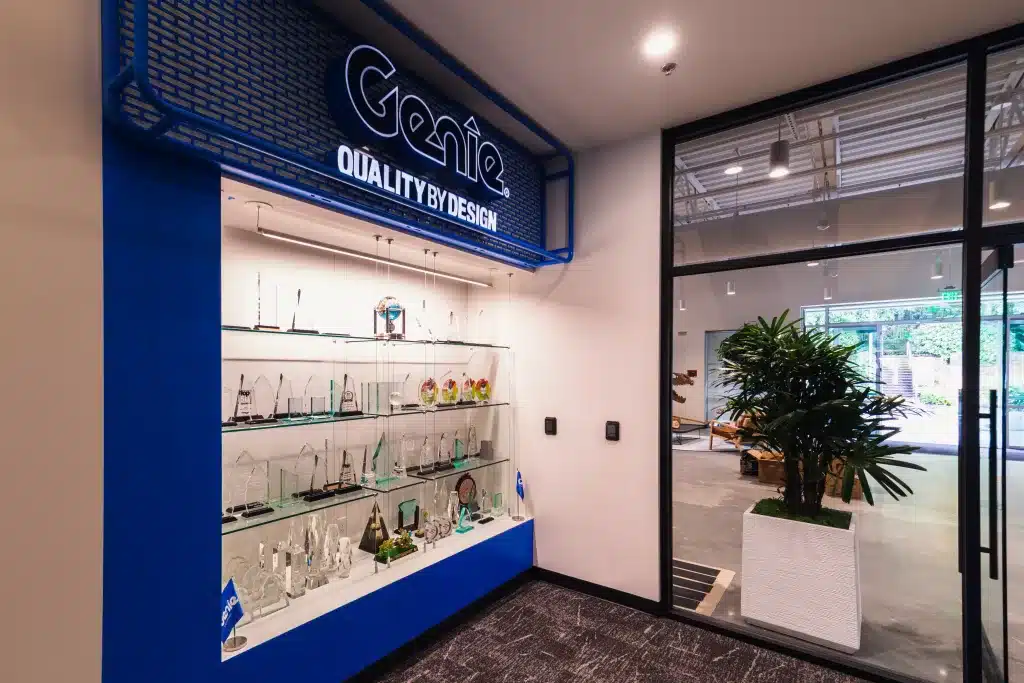 A display case with various awards and trophies in an office setting. A blue sign above reads "Genie Quality by Design." A large plant is visible next to a glass wall.