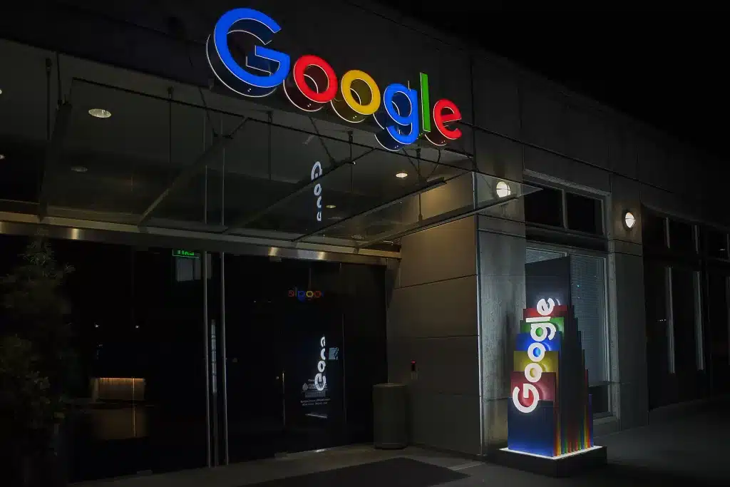Entrance to a Google office at night, featuring illuminated Google logos above the door and on a sign next to glass windows.