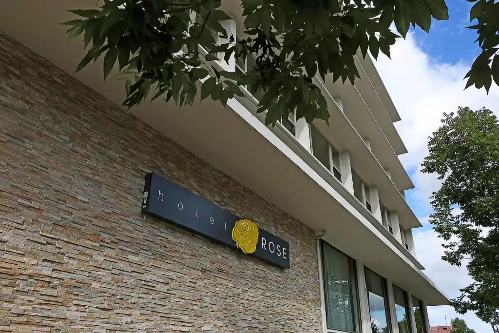 Exterior view of Hotel Rose, featuring a stone facade and a sign with a yellow rose logo, partially shaded by tree branches.