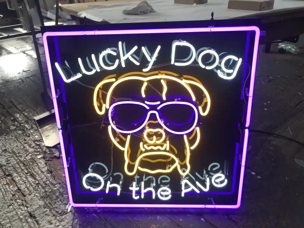 Neon sign displaying a stylized dog's face with sunglasses, surrounded by the text "Lucky Dog On the Ave" in white and yellow light.
