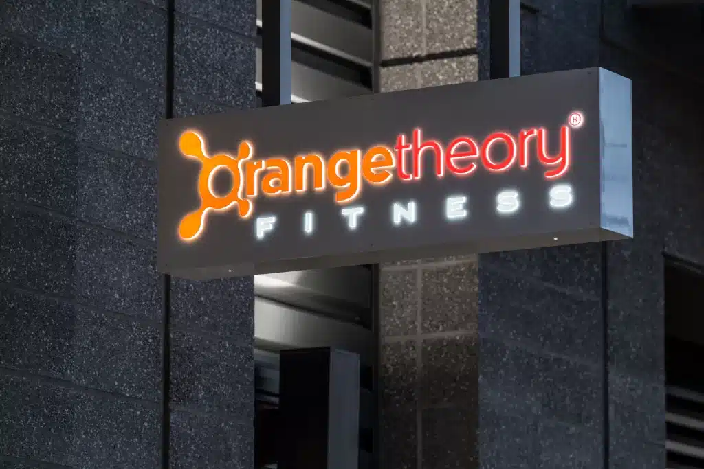 Sign for Orangetheory Fitness on a building exterior, featuring the gym's logo and name illuminated against a dark background.