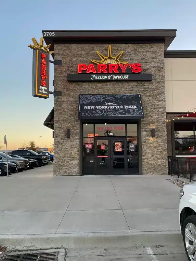 Exterior of Parry’s Pizzeria & Taphouse with a stone facade, illuminated signs, and advertisement for New York-style pizza. Parking lot and part of another building are visible.