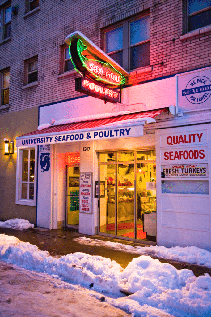 Street view of University Seafood & Poultry storefront at dusk, with neon signs and snow on the sidewalk.