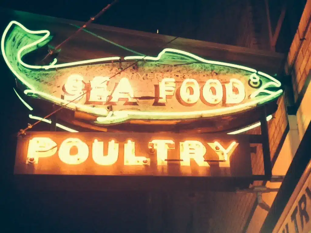 Neon sign shaped like a fish, displaying the words "SEA FOOD POULTRY" in glowing letters against a dark background.