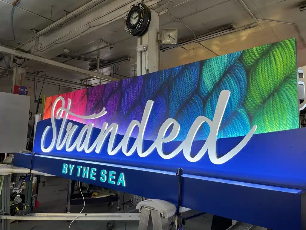 A colorful sign reading "Stranded by the Sea" in a workshop setting, featuring a rope texture background and bold, illuminated lettering.