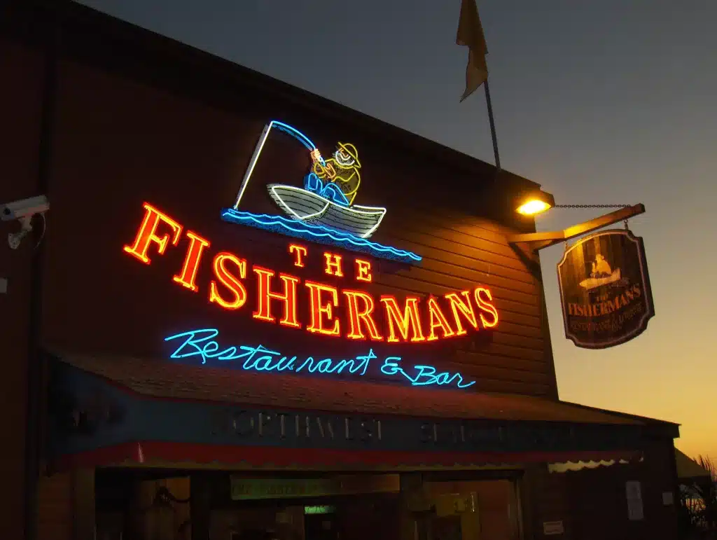 A restaurant exterior with a neon sign displaying "The Fisherman's Restaurant & Bar" featuring an illustration of a person fishing from a boat. The scene is taken at dusk.