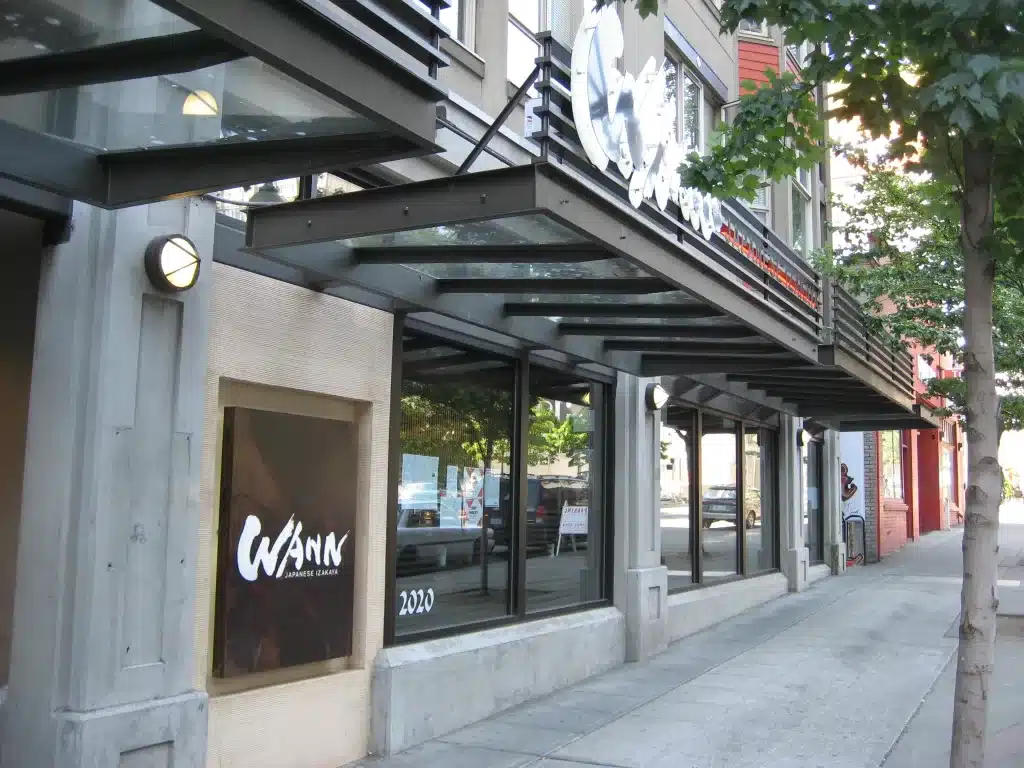 Street view of a modern building with large windows and a sign for "WANN Japanese Izakaya" on the facade. Trees line the sidewalk.