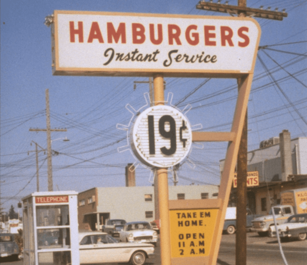 Vintage hamburger sign advertising "Instant Service" and hamburgers for 19 cents. A small notice reads "Take Em Home, Open 11 A.M. - 2 A.M." Surrounding area shows cars and a phone booth.