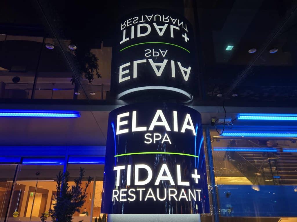 Illuminated sign with "ELAIA SPA" and "TIDAL+ RESTAURANT," reflecting on a glass surface, surrounded by blue ambient lighting.