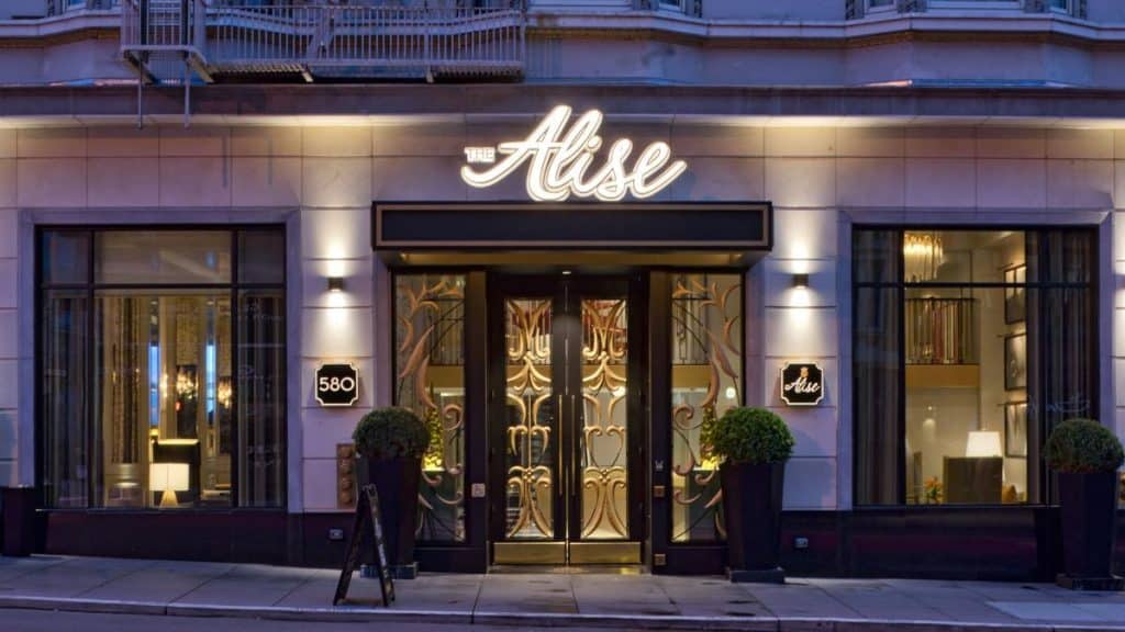 Street view of The Alise hotel entrance with illuminated sign, large windows on either side, potted plants, and a decorative glass door.