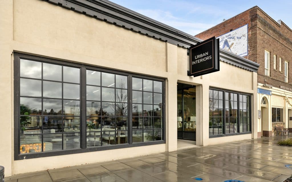 Storefront of Urban Interiors with large windows and a black sign. The exterior features beige walls and a sidewalk with a wet appearance.